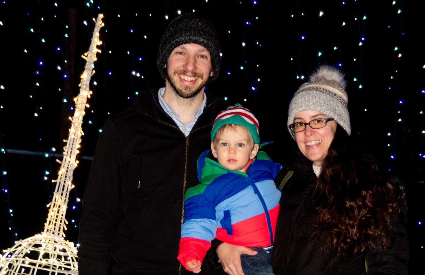 Husband and wife standing next to each other holding toddler in front of lights