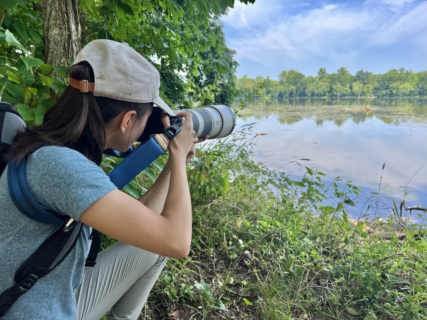 a girl squatting by the water looking through a camera with a large zoom lens