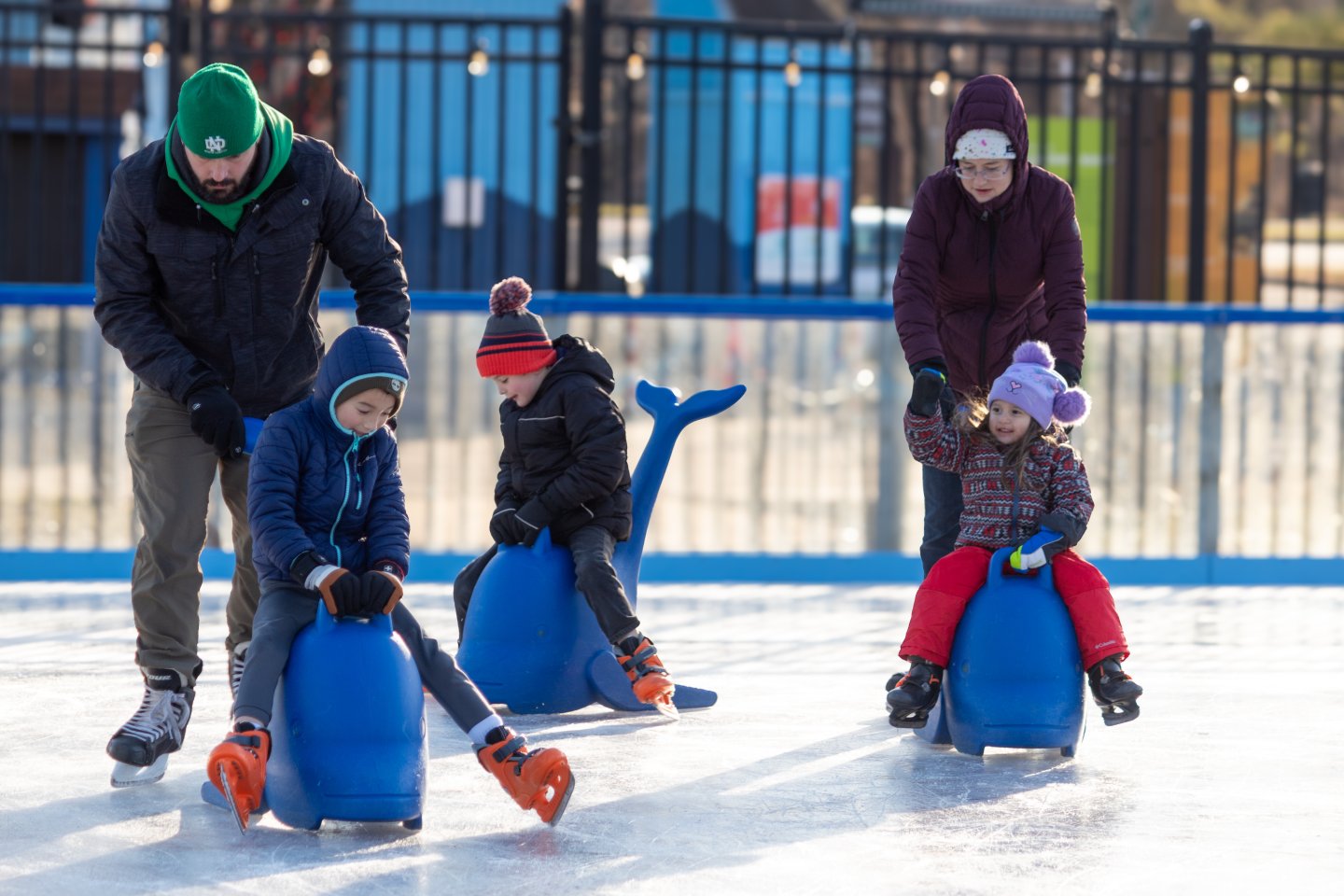 A family ice skating at Cameron Run. Three children riding on top of penguin ice skating helpers.