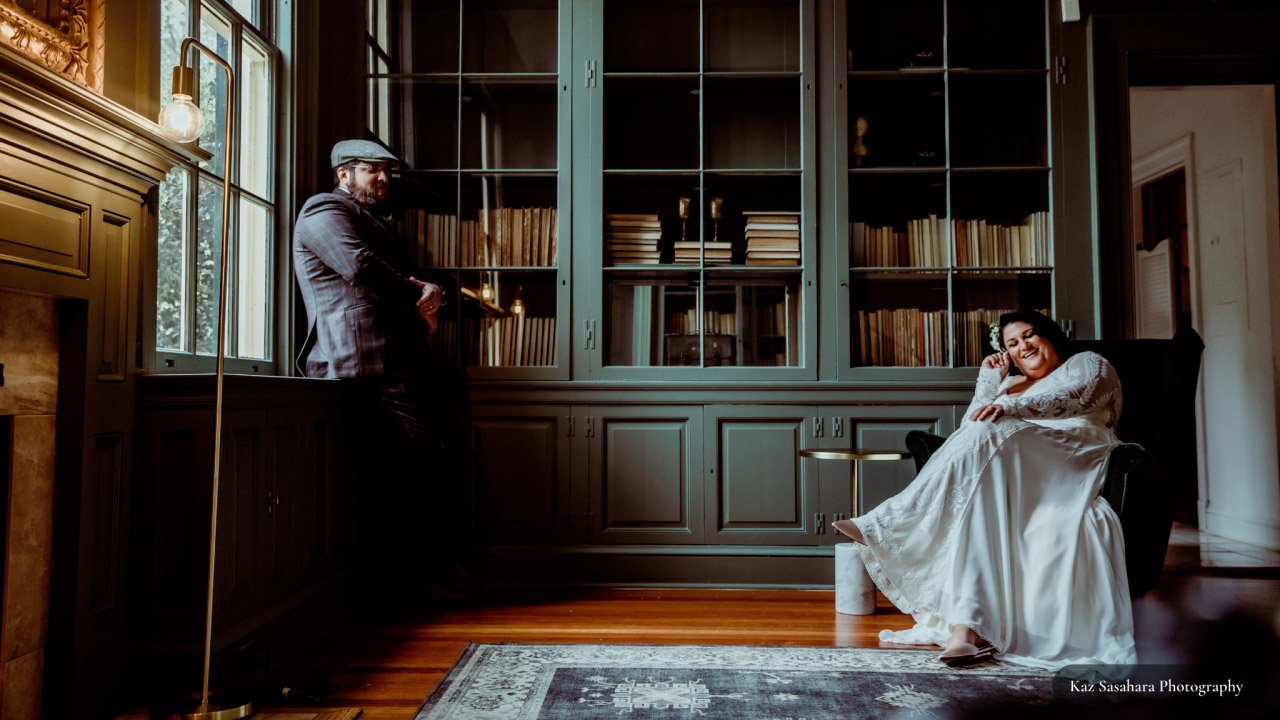 Bride and groom in darkened Rust Manor House library bride is sitting in a chair