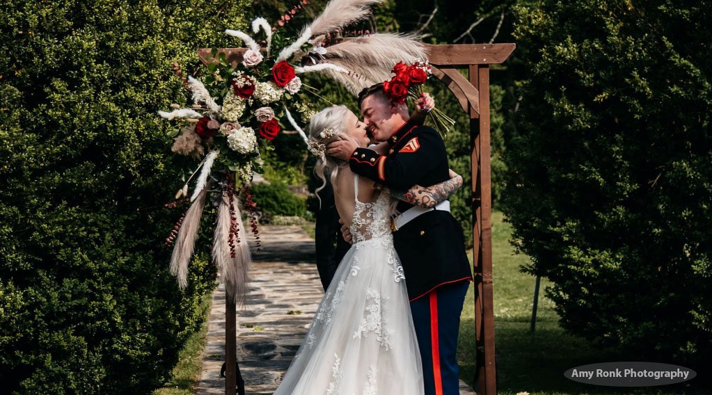 Bride and groom in front of pergola. Groom wearing military uniform. Red and white  flowers on pergola.