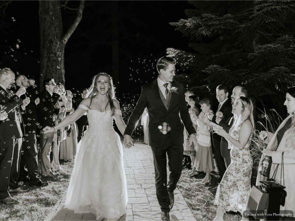 Black and white image of bride and groom leaving wedding reception with guests congratulating them
