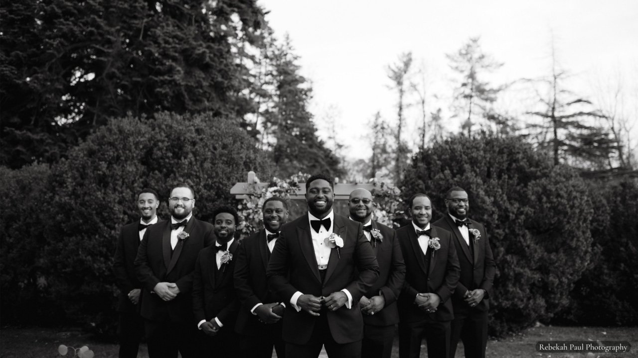 Black and white image of grooms party in tuxedos in front of conifer trees