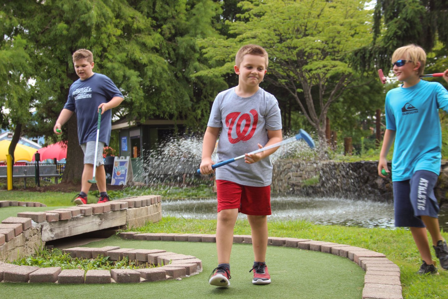 Three children playing mini-golf on a course with a water feature in the background. One child is in the foreground, swinging a golf club, with two others watching in the background.