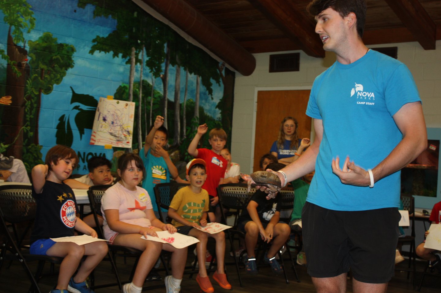 An educator from NOVA Camp River wearing a blue shirt holds a small turtle, presenting to seated children in a room with a forest mural on the wall.
