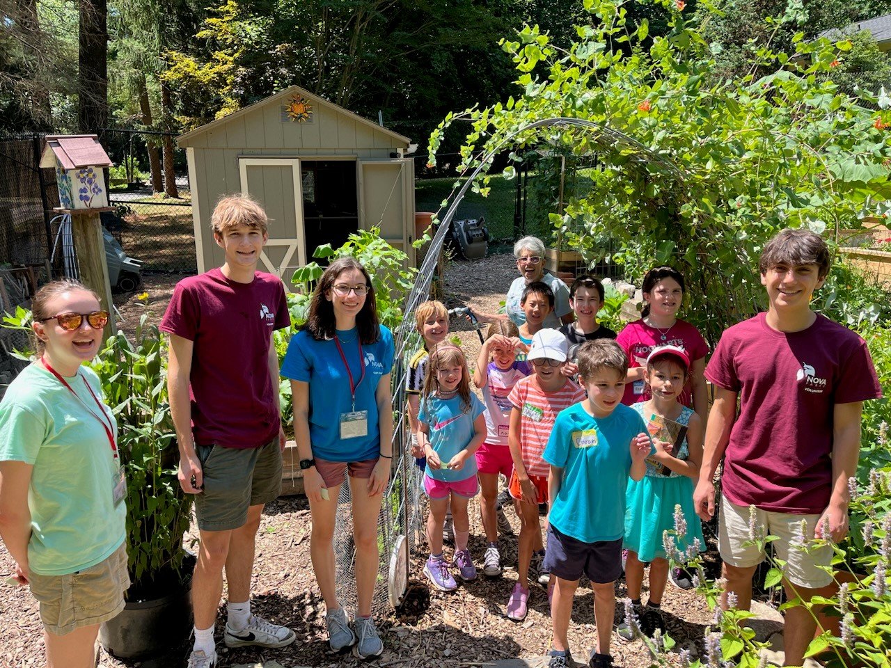 A group of children and teenagers posing outside in a garden, with a shed and trees in the background, all wearing casual summer clothing.