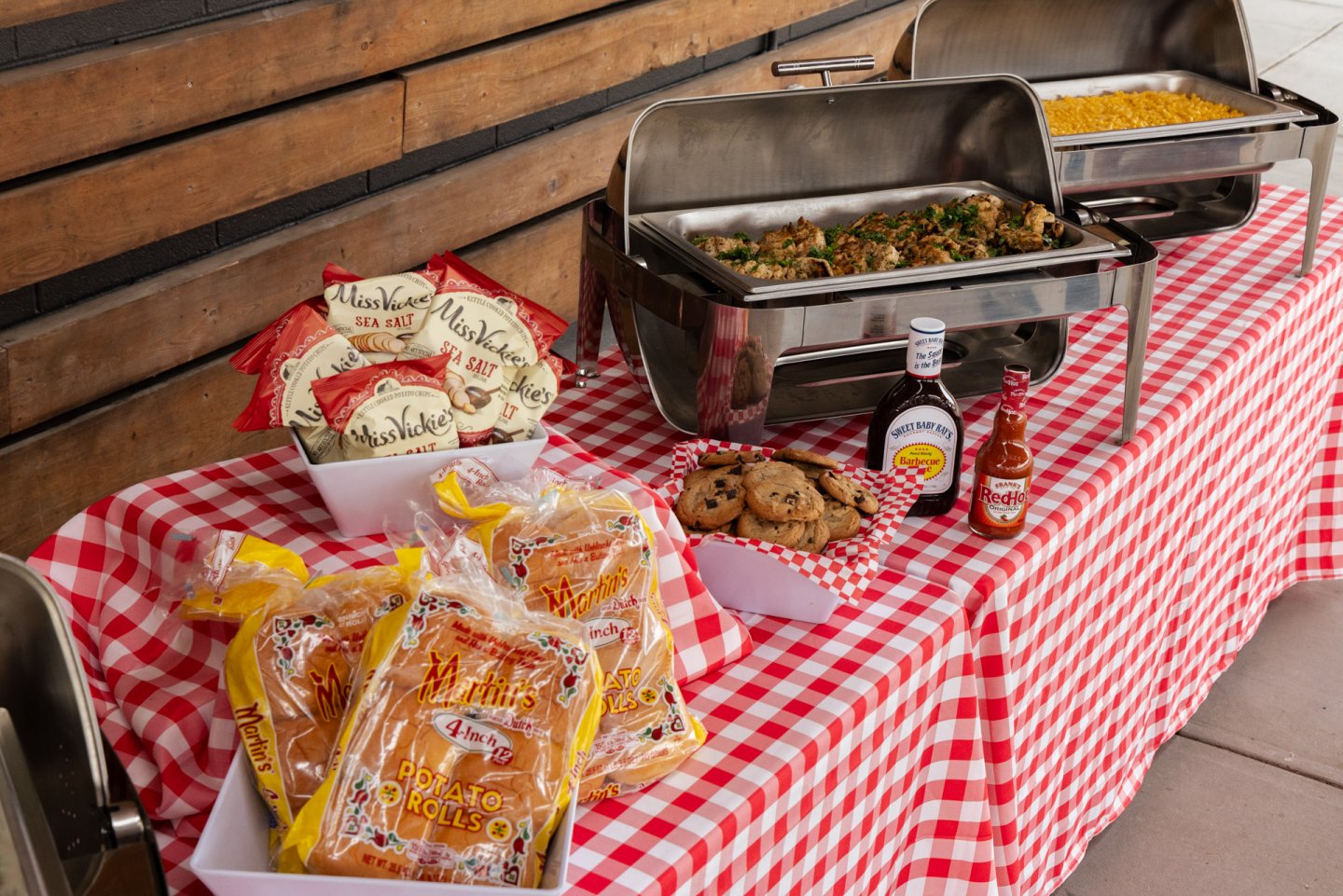 Buffet table with food trays, bread, cookies, and condiments on a red checkered cloth.