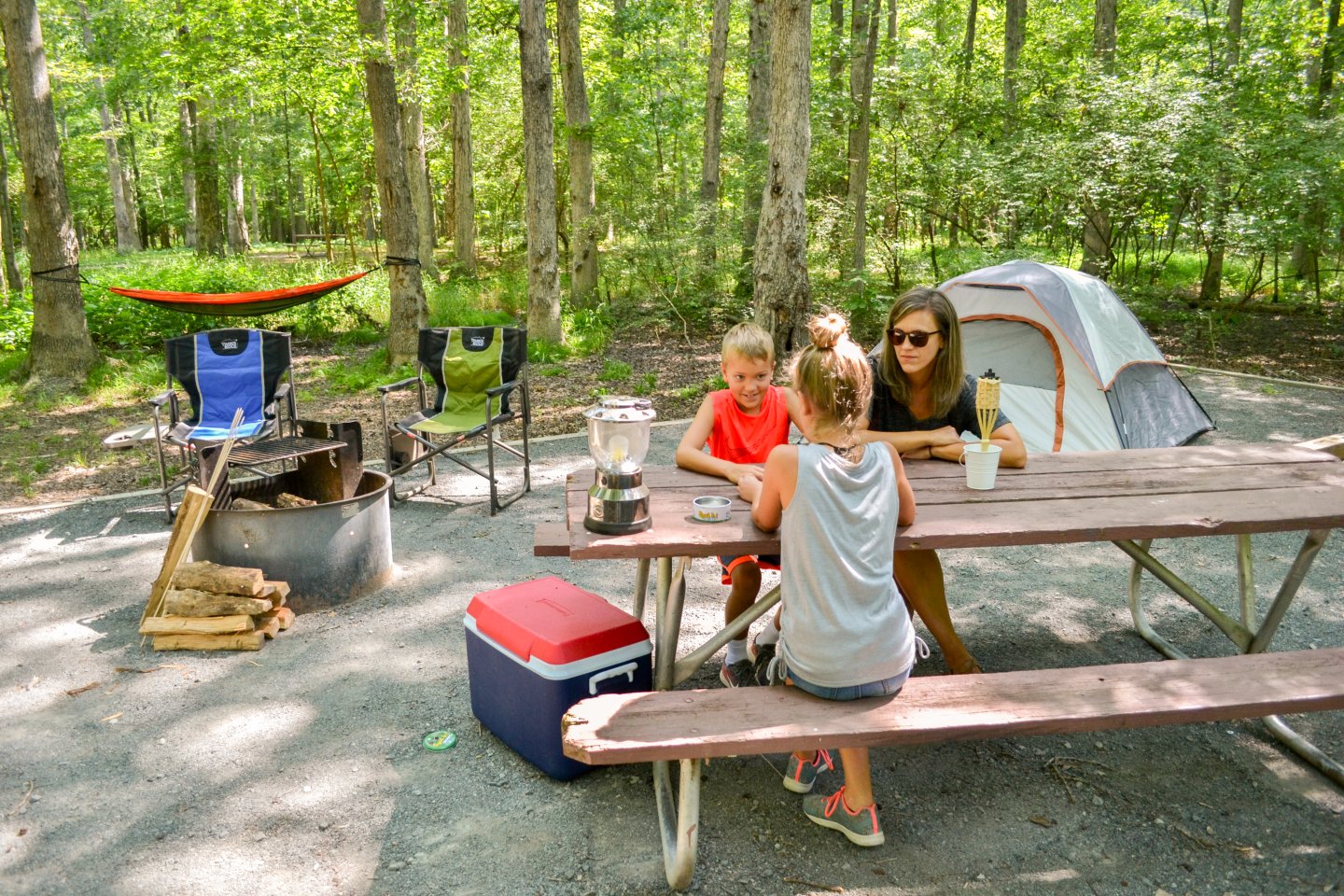 Family sitting at a picnic table in a forest campsite with a tent nearby.