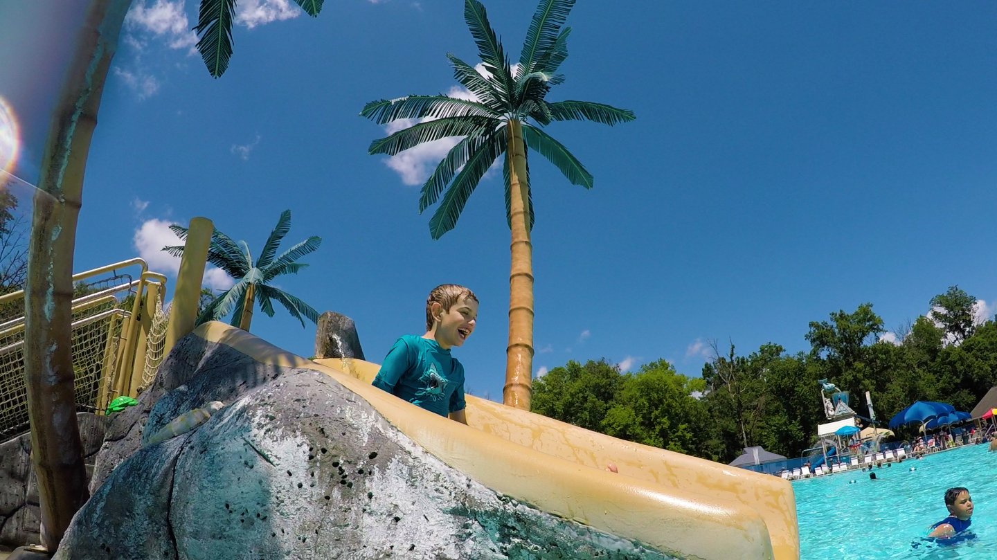 Child on water slide at a pool, with palm trees and a bright blue sky.