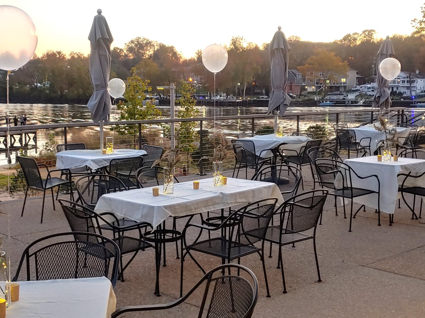 Outdoor restaurant tables by a riverside at sunset, decorated with lanterns and balloons.