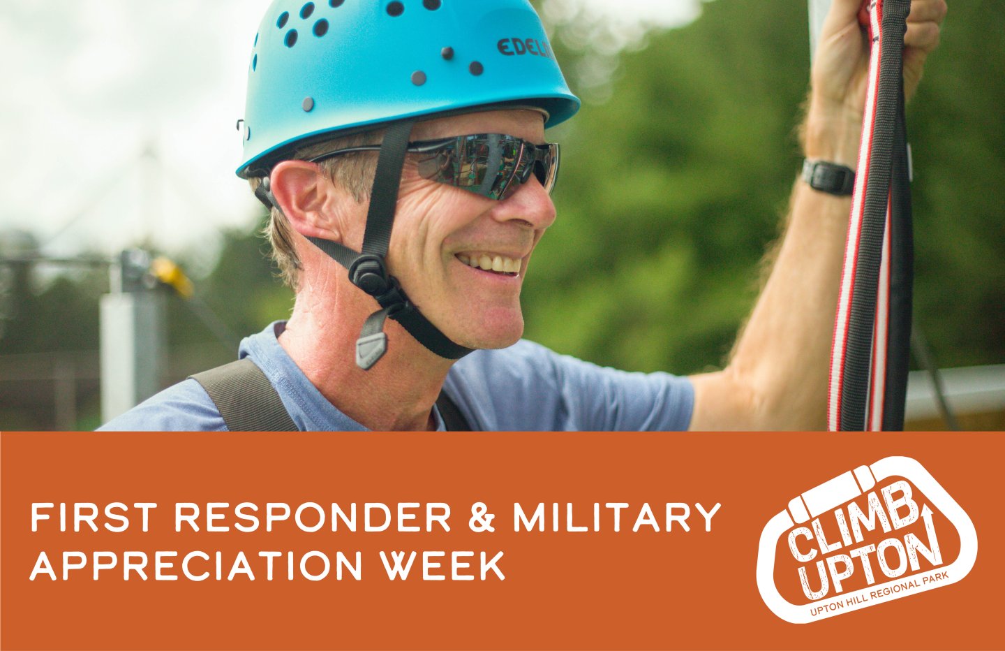 Man climbing with helmet and sunglasses, smiling outdoors. Climb Upton logo on the side.