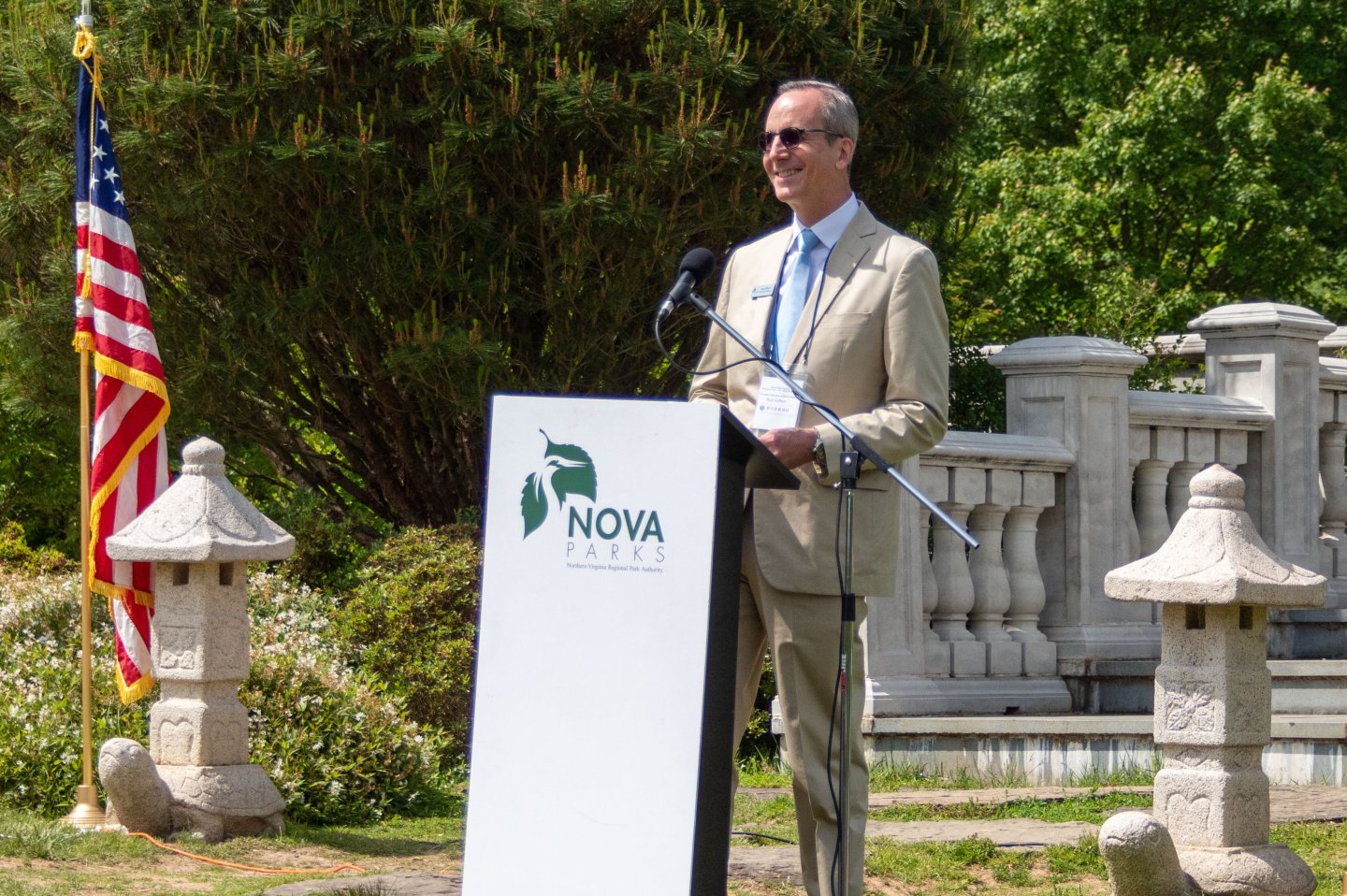 Man speaks at podium outdoors, U.S. flag nearby, sunny day.