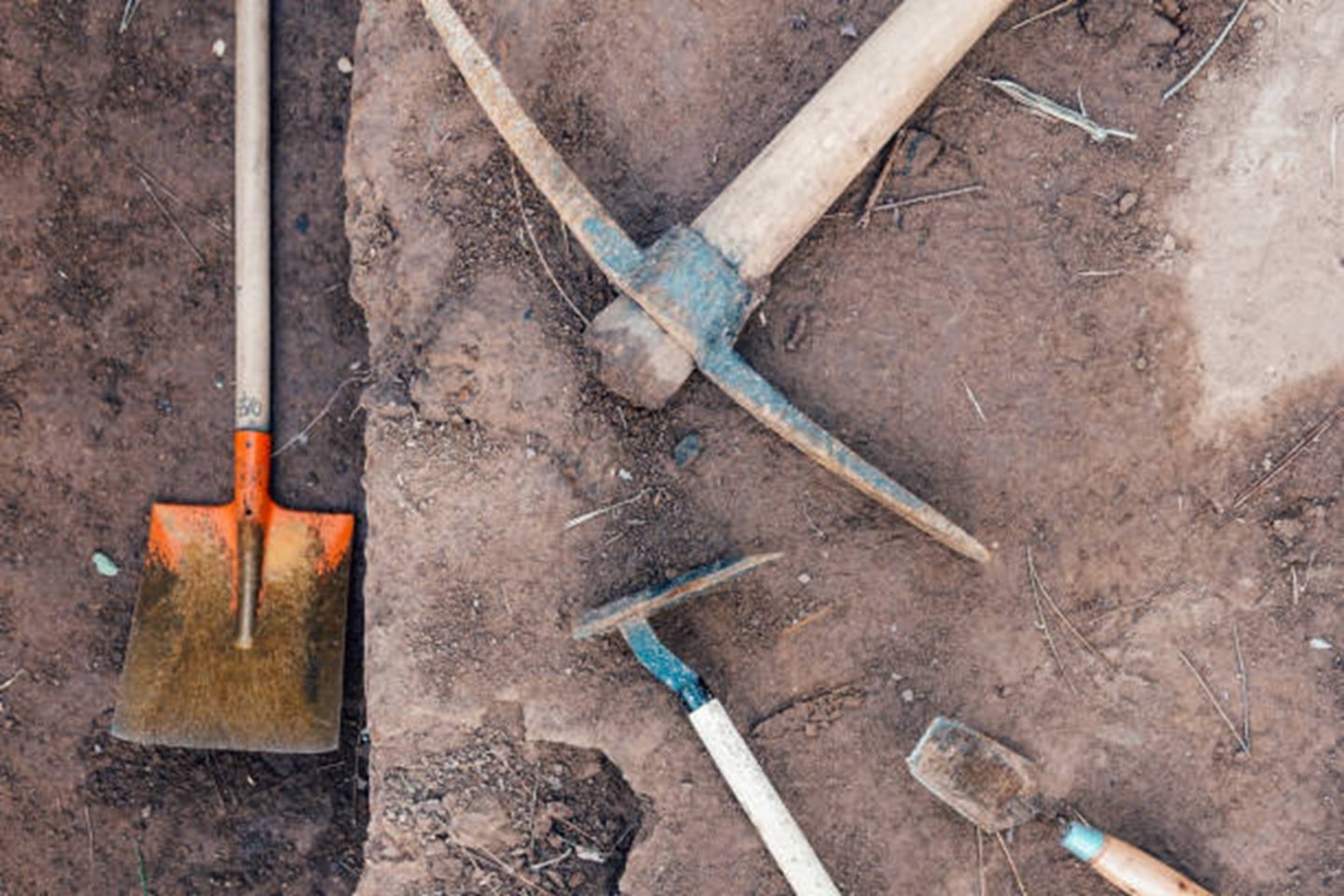 Shovel, pickaxe, and trowel on brown dirt ground.