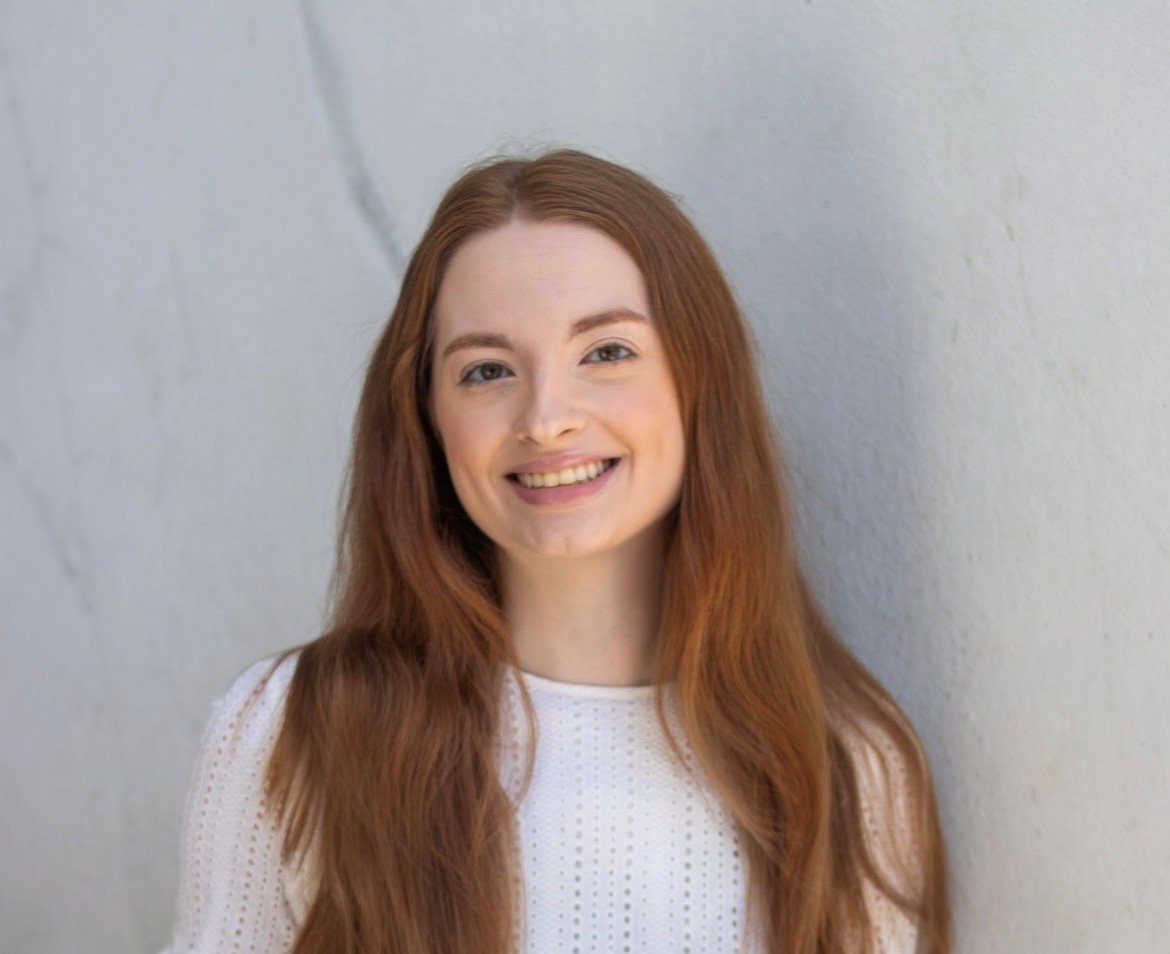 Smiling woman with long red hair against a light gray textured wall.