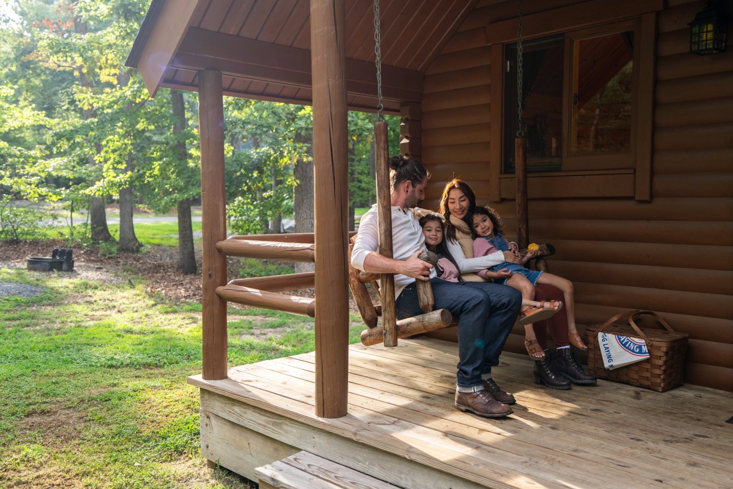 Family reading on a porch swing of a log cabin, surrounded by greenery.