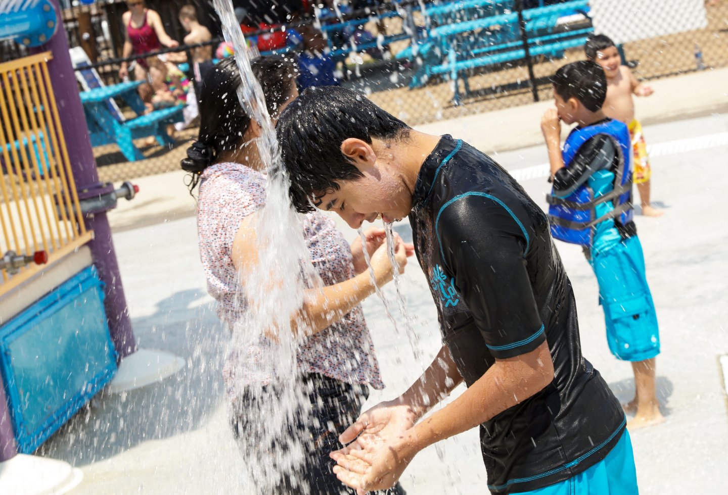 Boy in swimwear under a water shower at a park, people in the background.