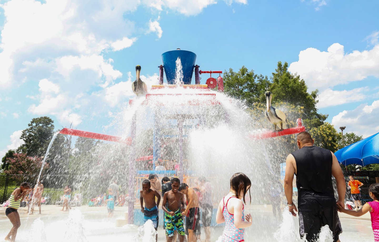 Kids and adults play under a water fountain at a splash park on a sunny day.