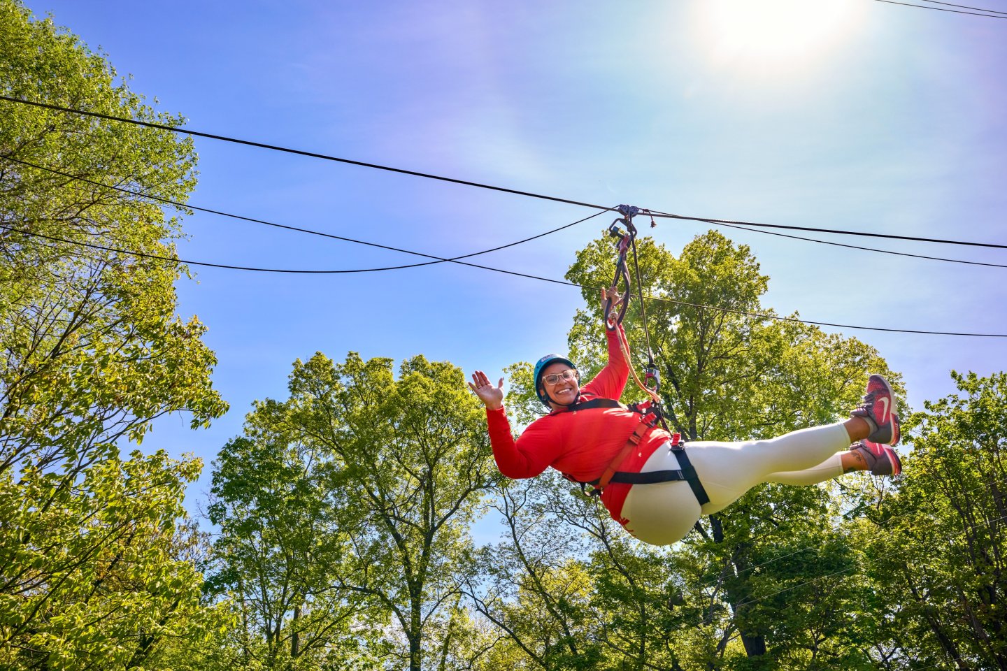 Person zip-lining through trees under a sunny sky, smiling and waving.