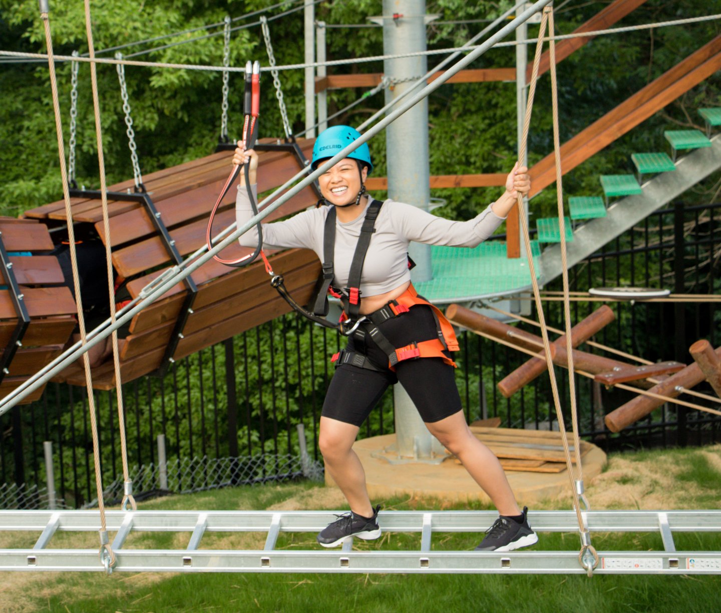 Woman on rope bridge smiling, wearing helmet and harness in adventure park.