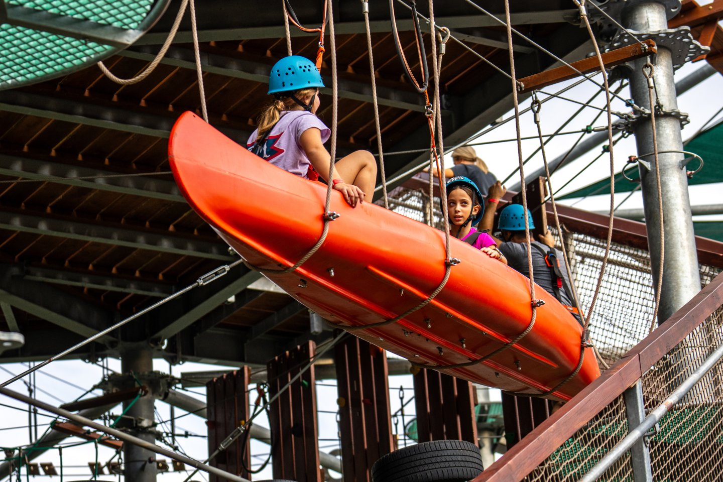 Kids wearing helmets on a ropes course in an orange kayak.