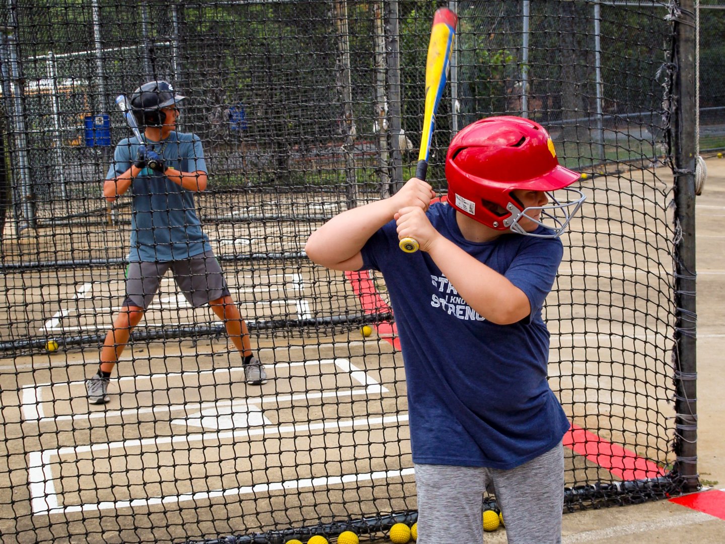 Boy and man practicing baseball in a batting cage, both wearing helmets.