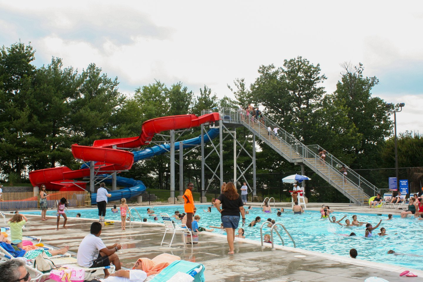 Outdoor pool with water slide, swimmers, and trees in the background.