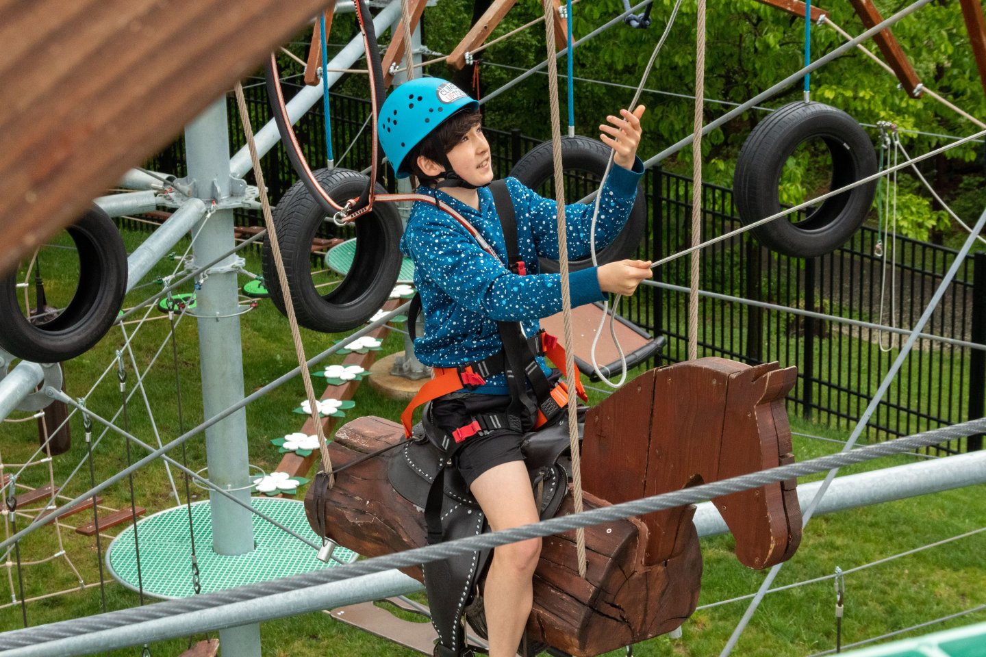 Person in a helmet on rope course riding a wooden horse, surrounded by greenery.