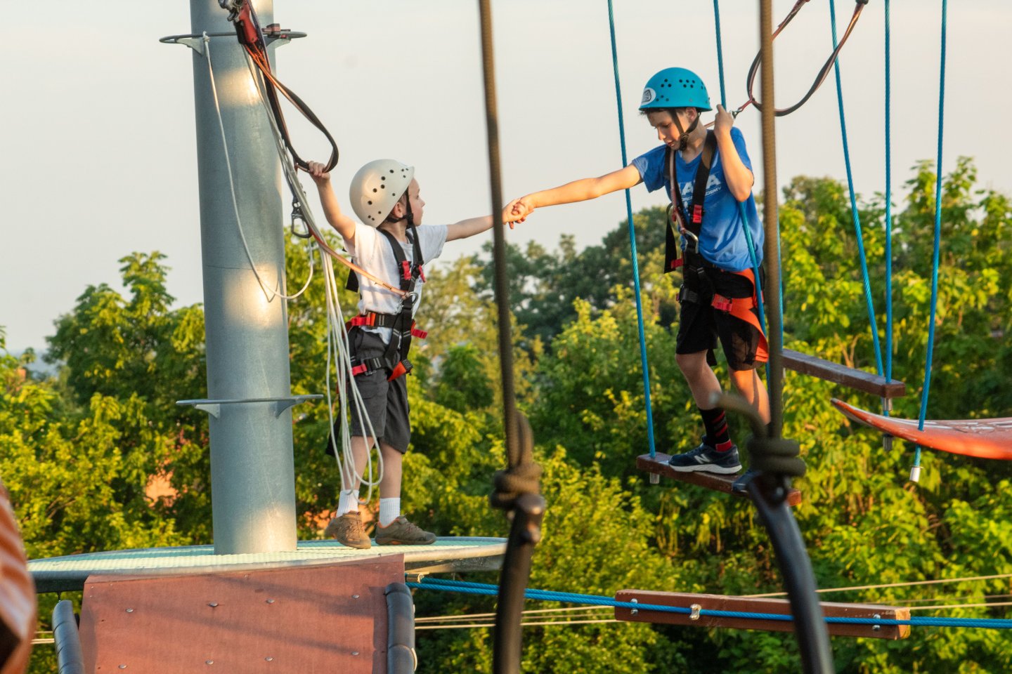Two children on a ropes course, wearing helmets, reaching out to each other.