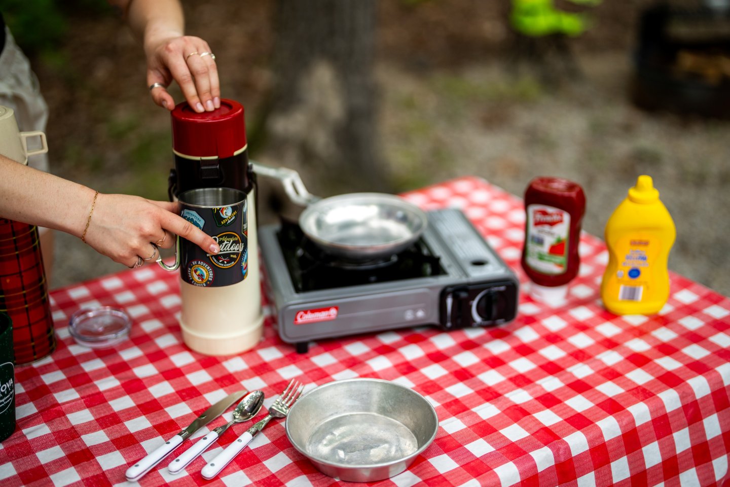Camping setup with red checkered tablecloth, burner, condiments, and a person's hands.