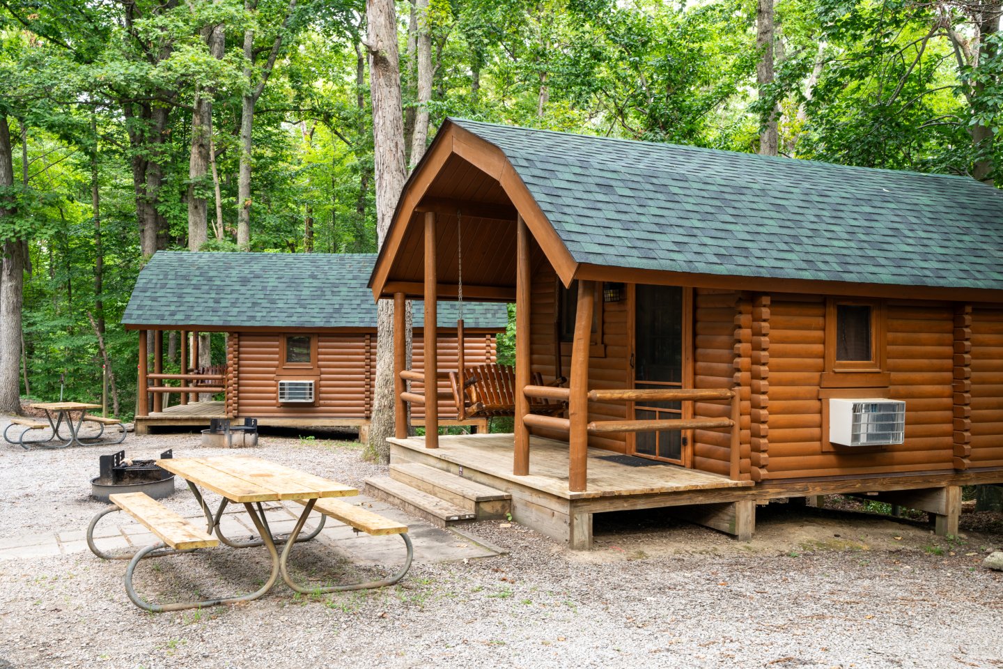 Two wooden cabins in a forest with picnic tables and a fire pit nearby.