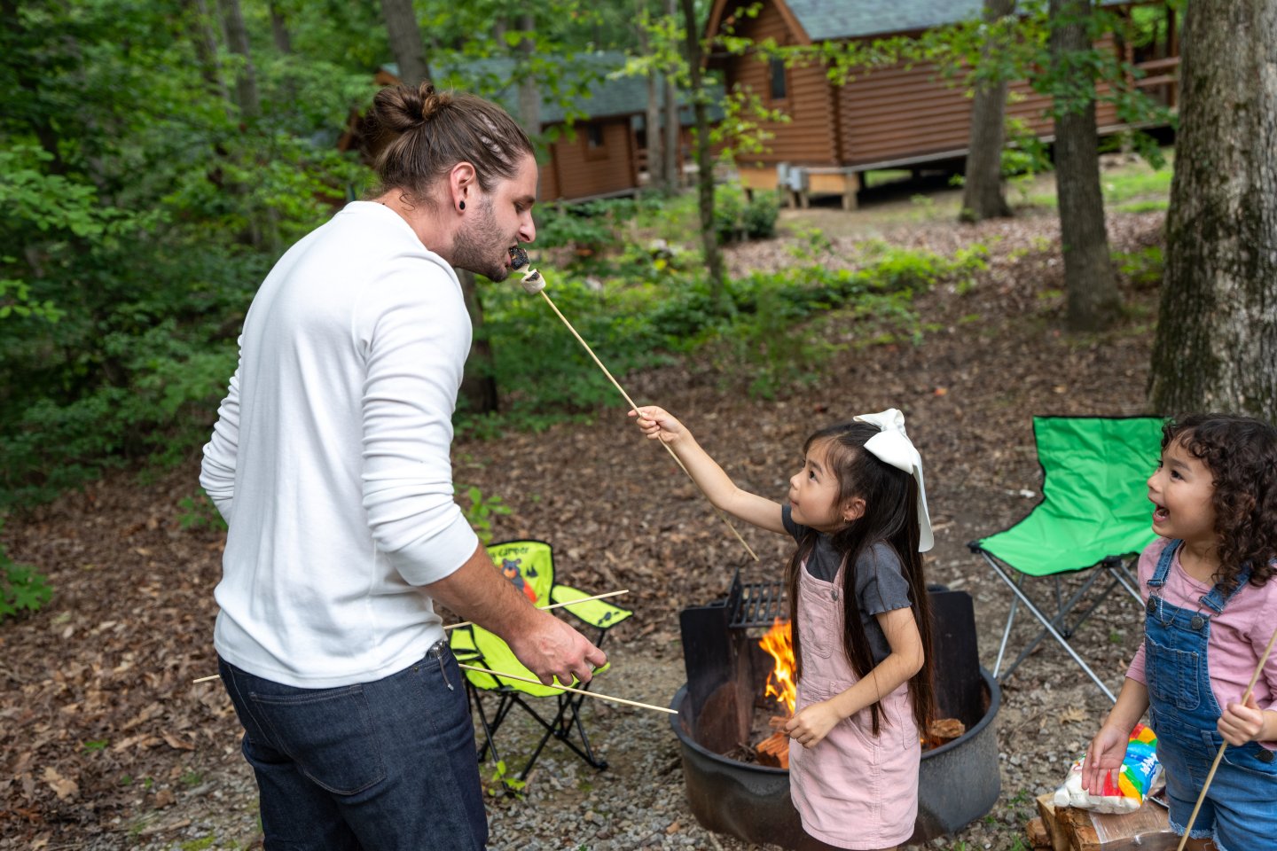 Man playfully toasting marshmallows with two smiling children at a campfire.