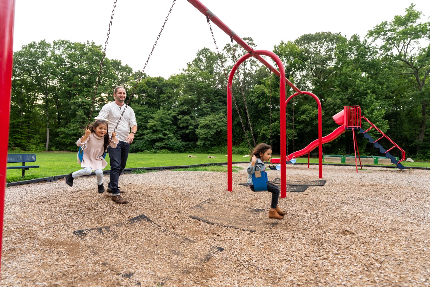 Children and adult playing on swings in a park, surrounded by trees.