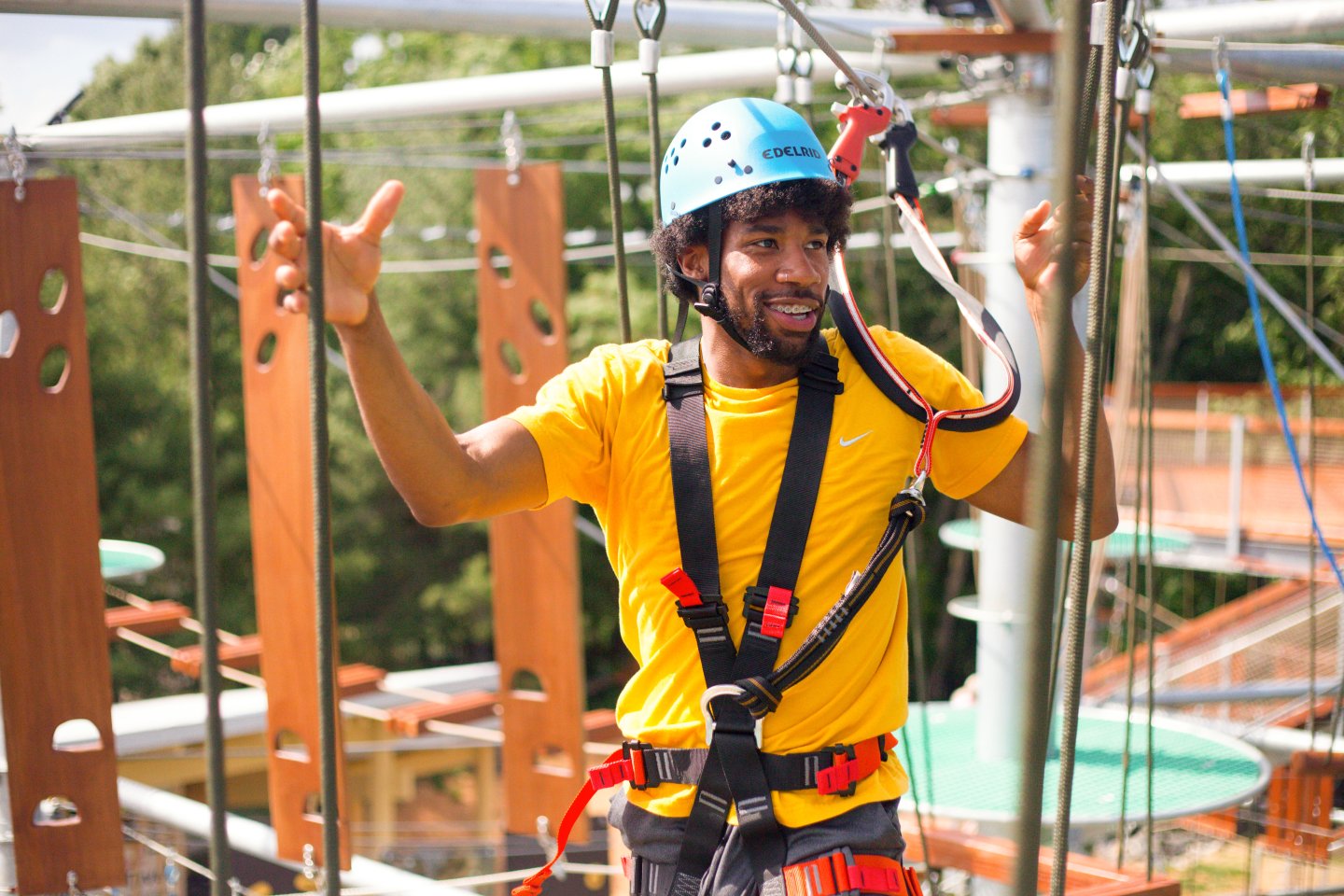 Man in a yellow shirt smiling on a ropes course, wearing a helmet and harness.