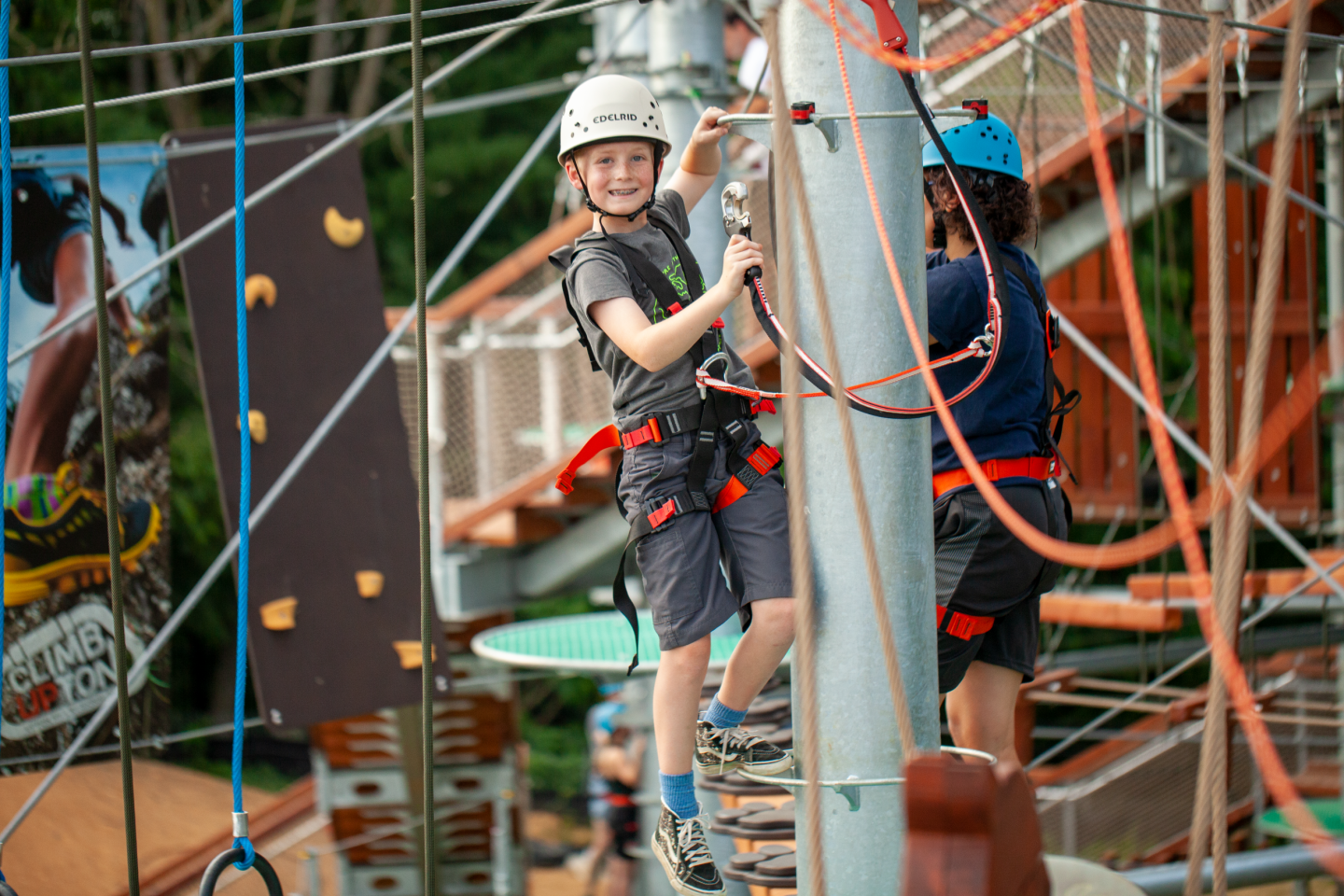 Kids in helmets climbing a rope course, smiling and wearing harnesses.