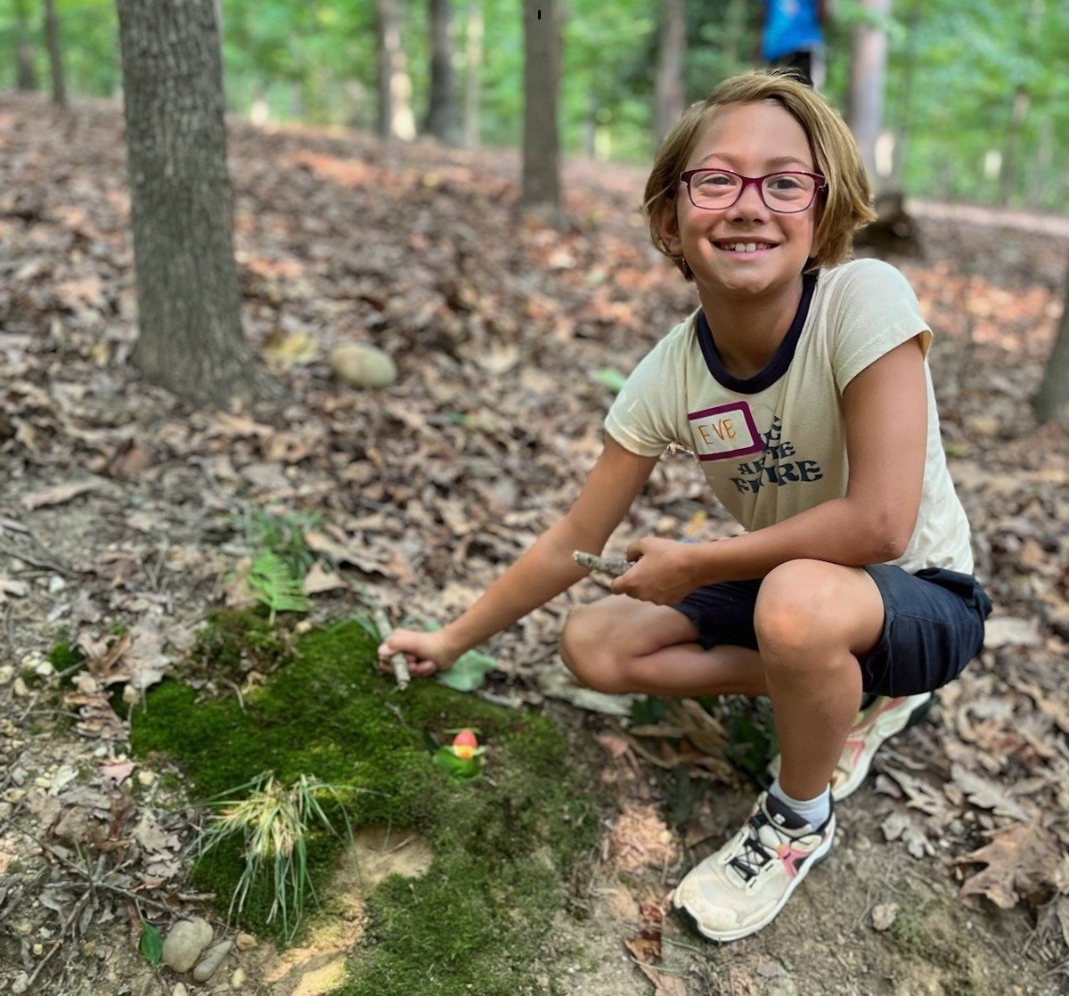 Smiling child crouches in a forest, arranging moss on the ground.