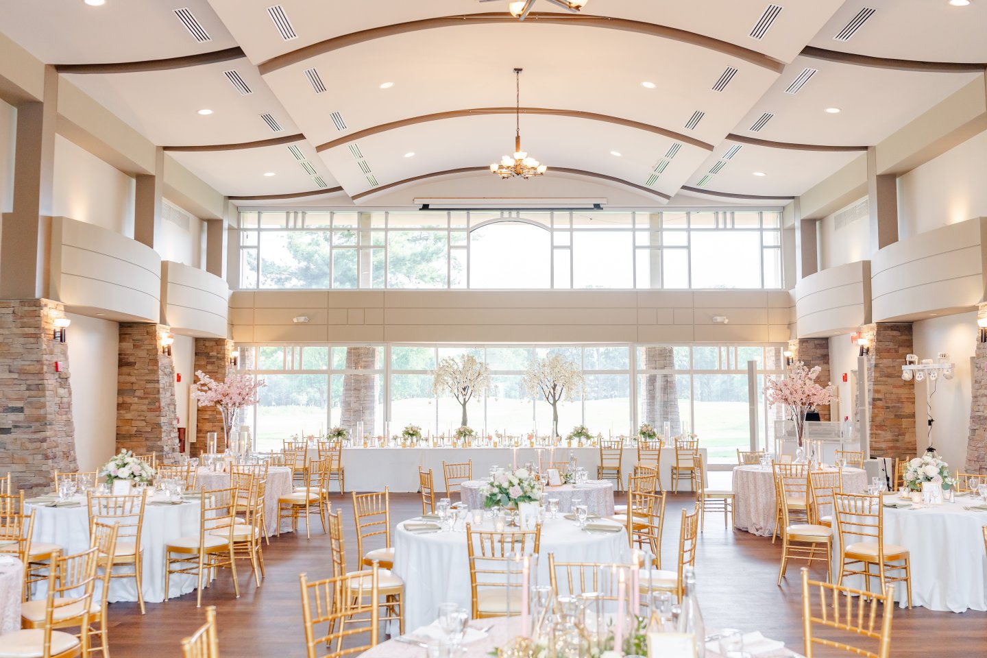 Bright event space with large windows, decorated for an event with white and gold chairs.