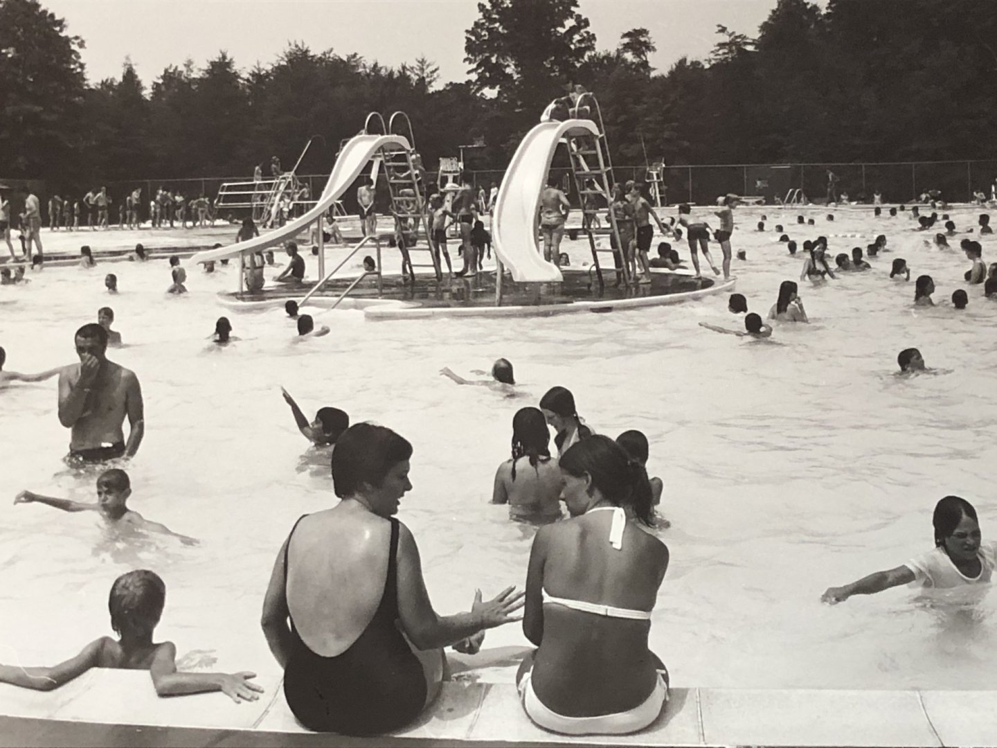 Crowded swimming pool with two slides, people swimming and sitting at the edge.