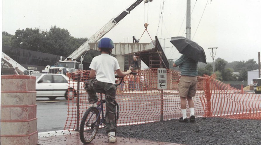 Boy on bike and man with umbrella watch crane at construction site.