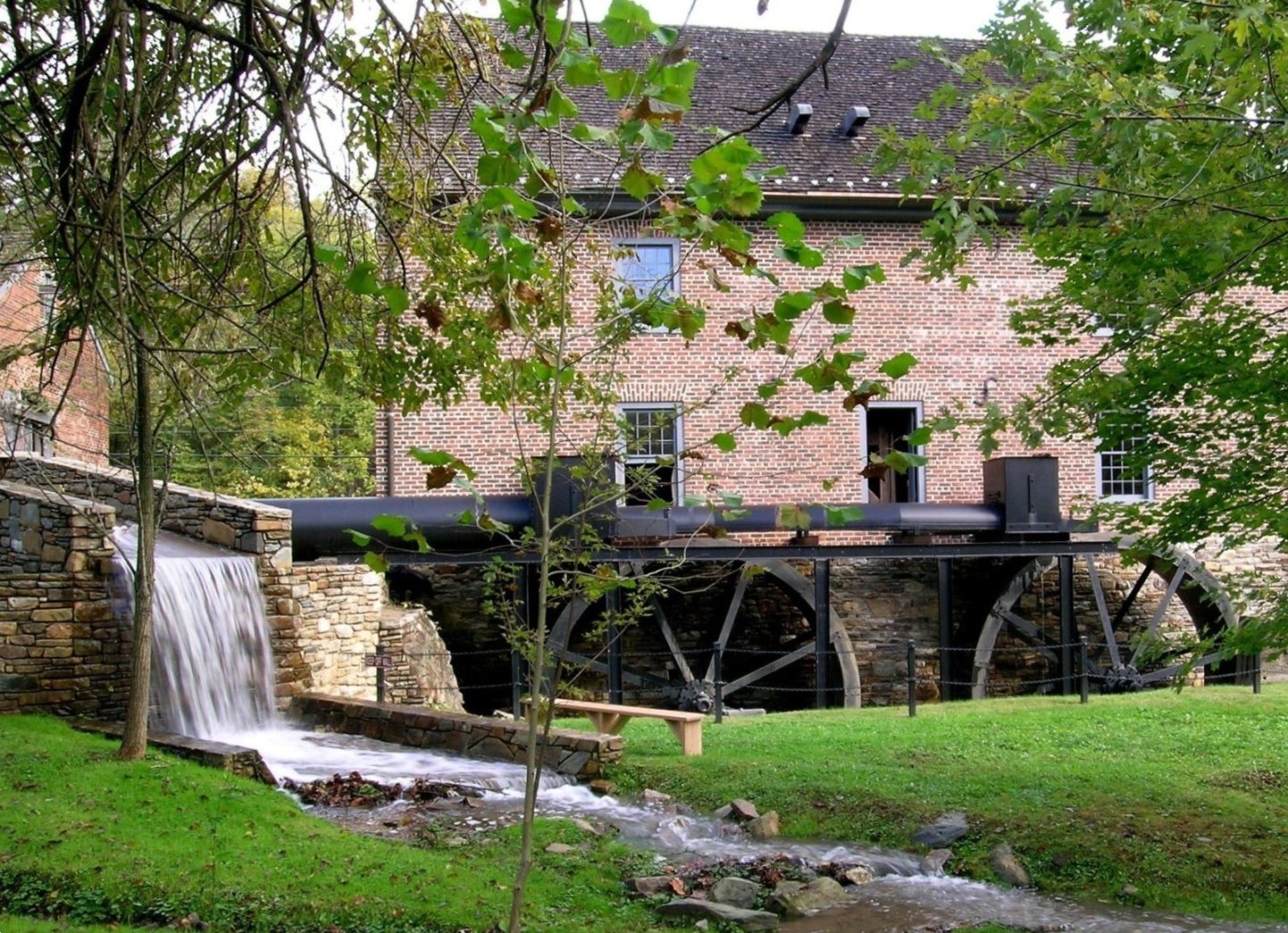 Brick watermill with large wheel and flowing stream, surrounded by trees.