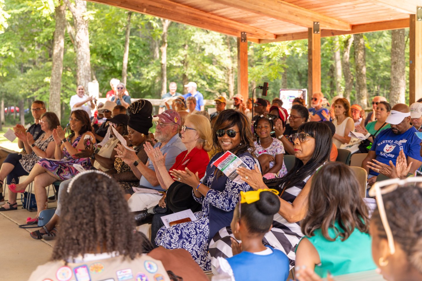 Diverse group seated under a pavilion in a park, clapping and smiling.