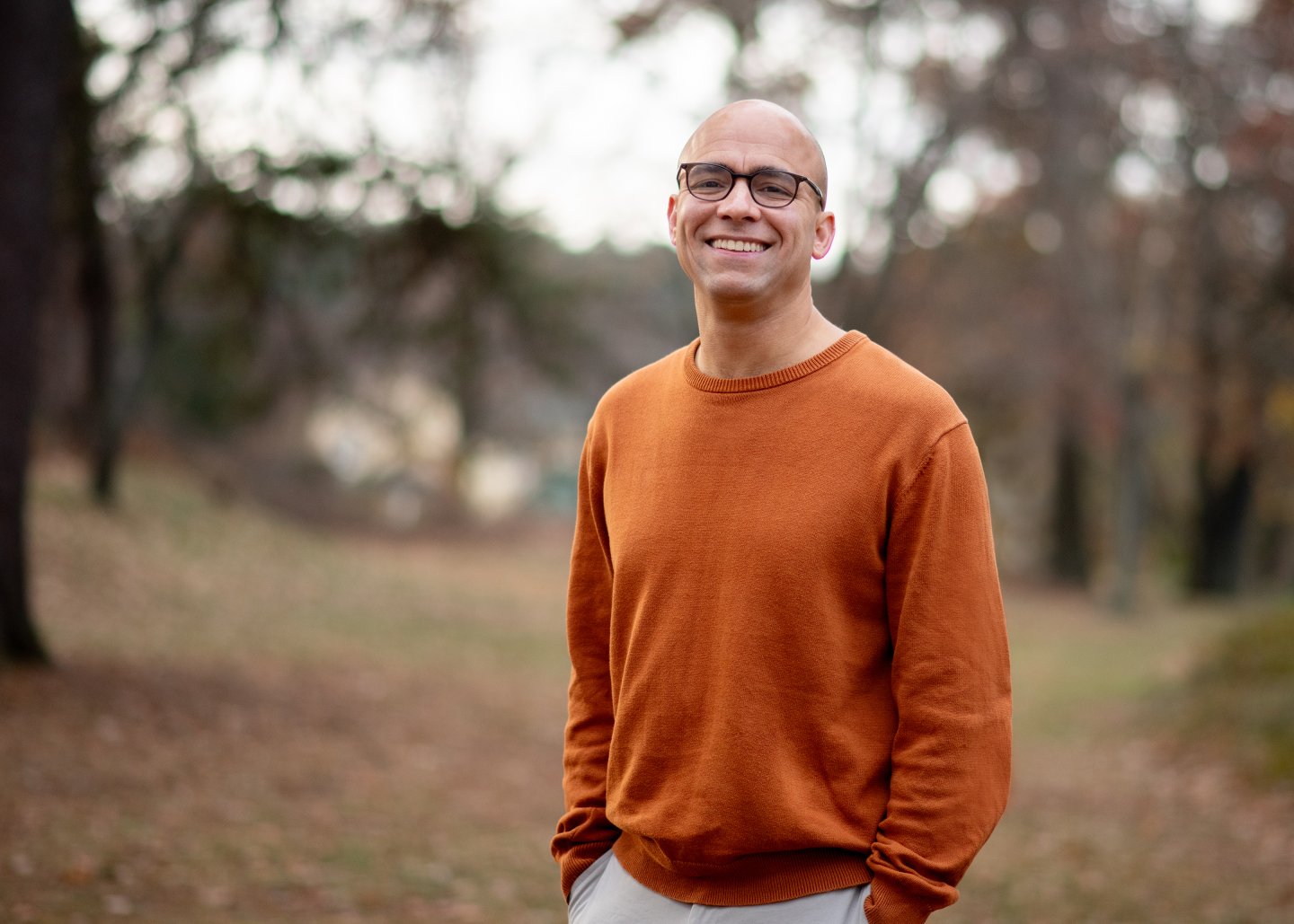 Smiling man in an orange sweater stands in a park with autumn foliage.