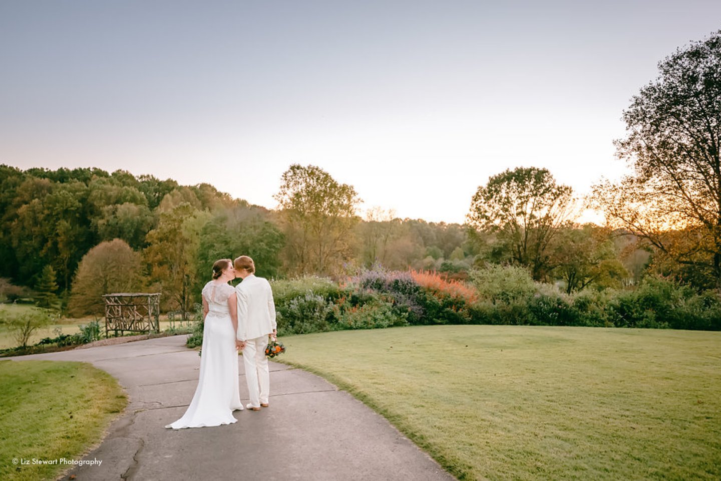 A couple in wedding attire walks down a garden path at sunset.