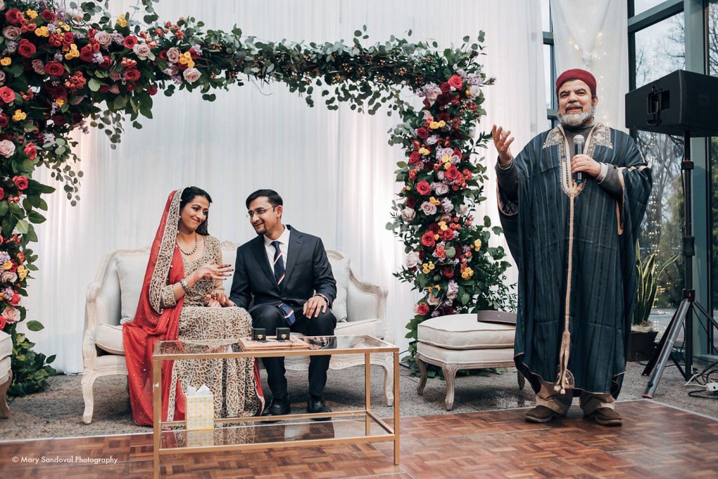 Bride and groom sit under floral arch with a speaker beside them.
