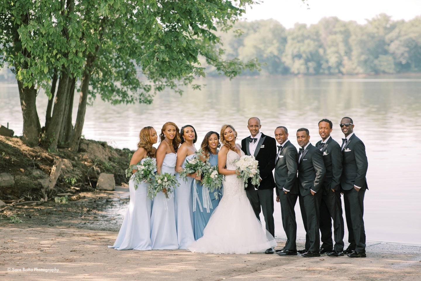 Wedding party by a lake, bridesmaids in light blue, groomsmen in dark suits.
