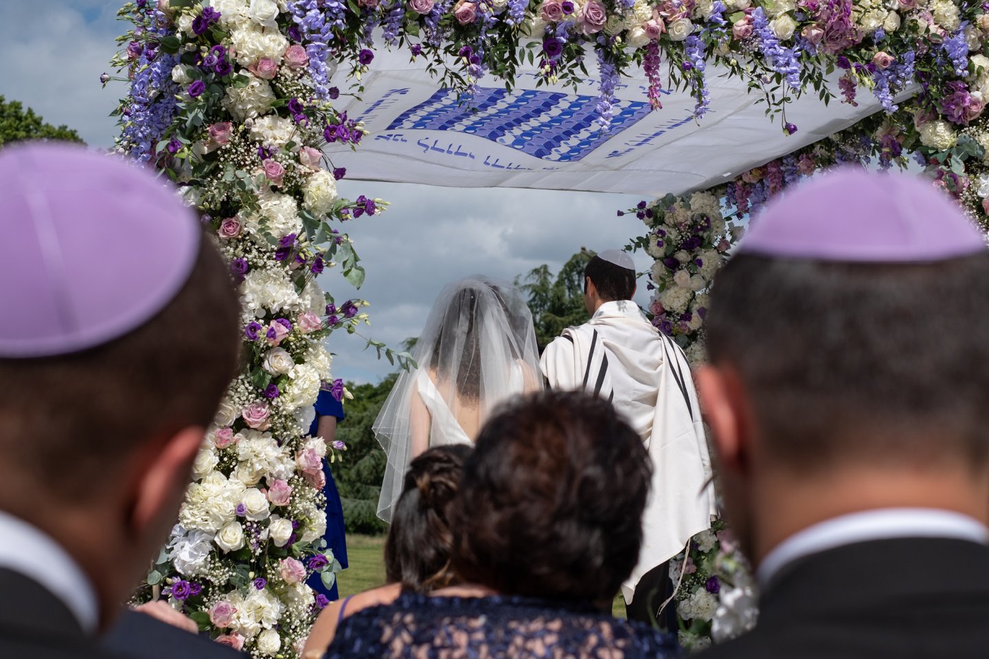 Outdoor wedding ceremony under floral canopy, bride and groom facing rabbi.
