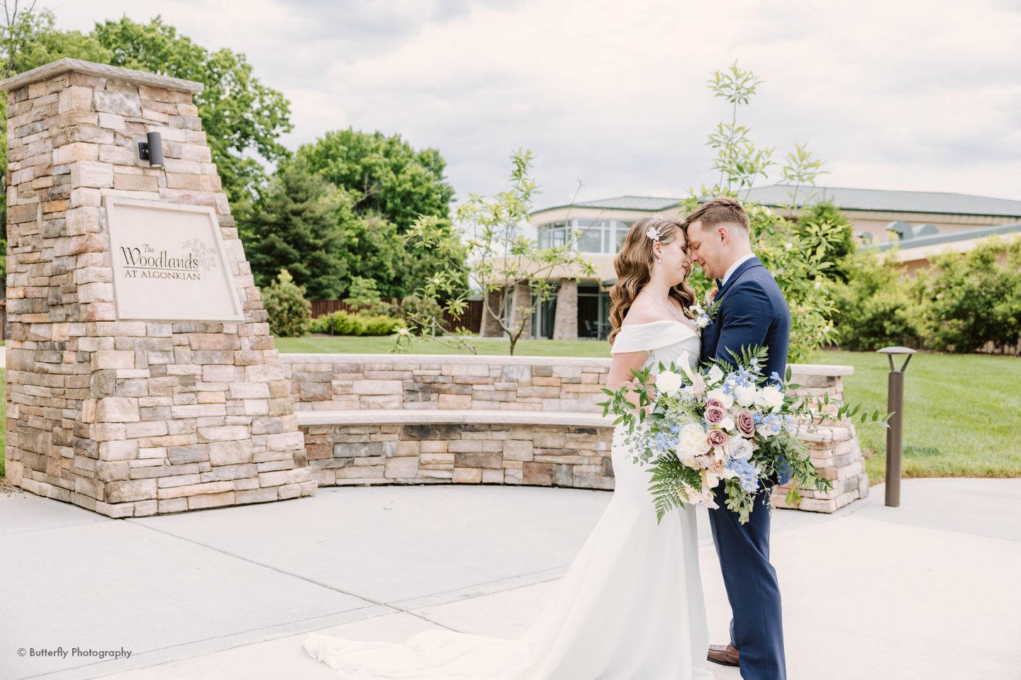 Couple embraces outdoors near stone wall, holding a bouquet.