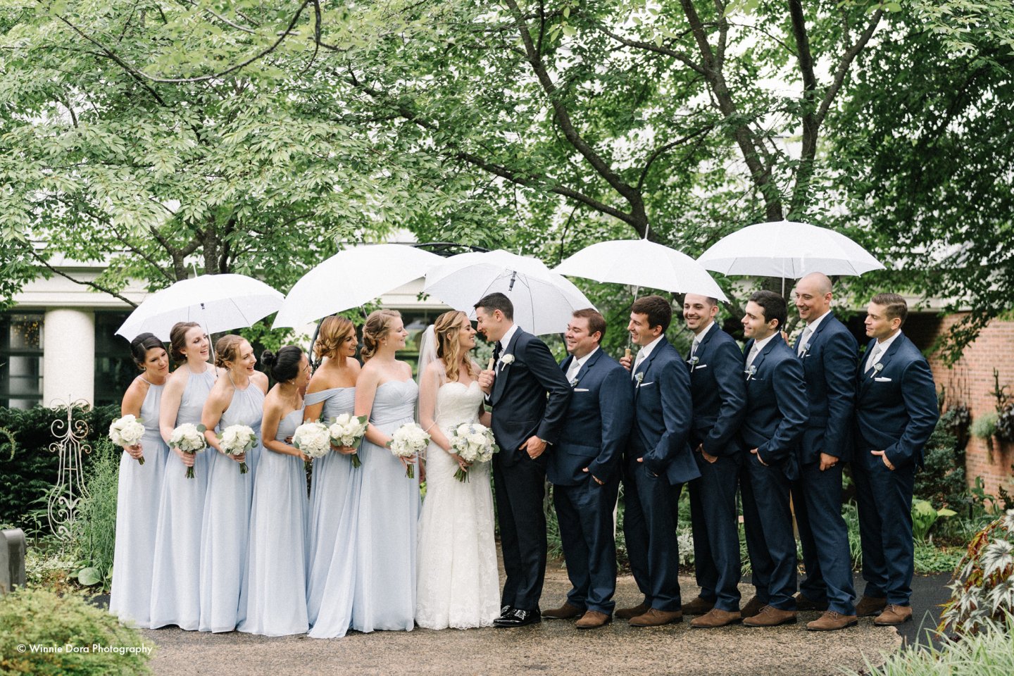 Bridal party with white umbrellas in a lush garden.