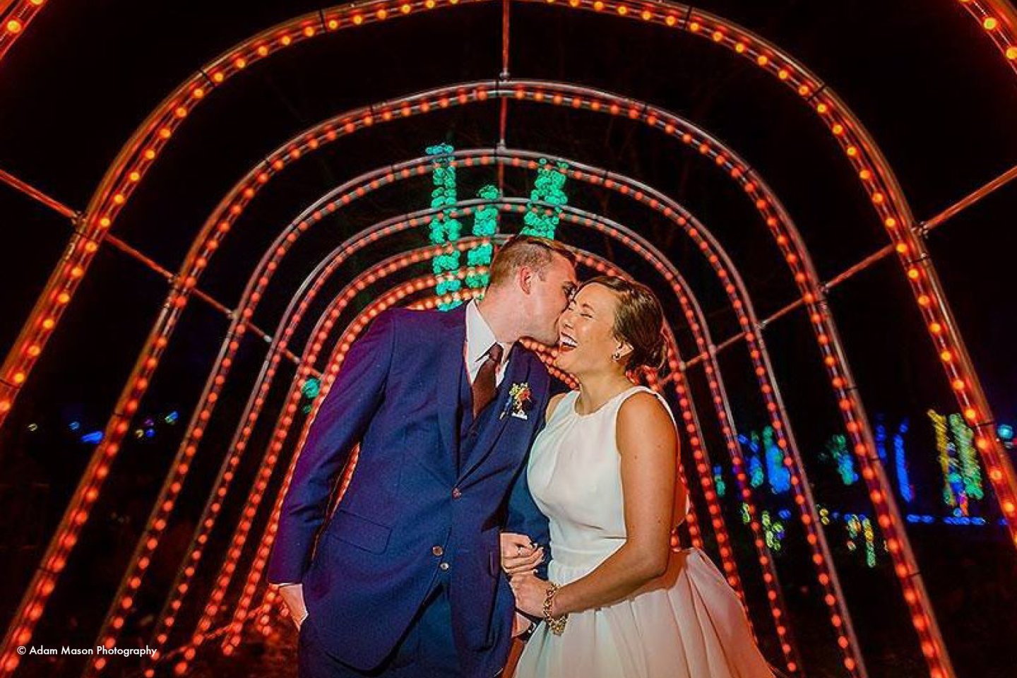 Couple in wedding attire under illuminated archway at night, smiling at each other.