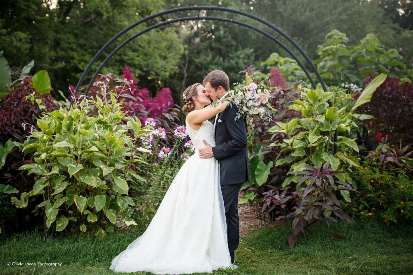 Bride and groom kissing in a garden under an arch, surrounded by vibrant greenery.