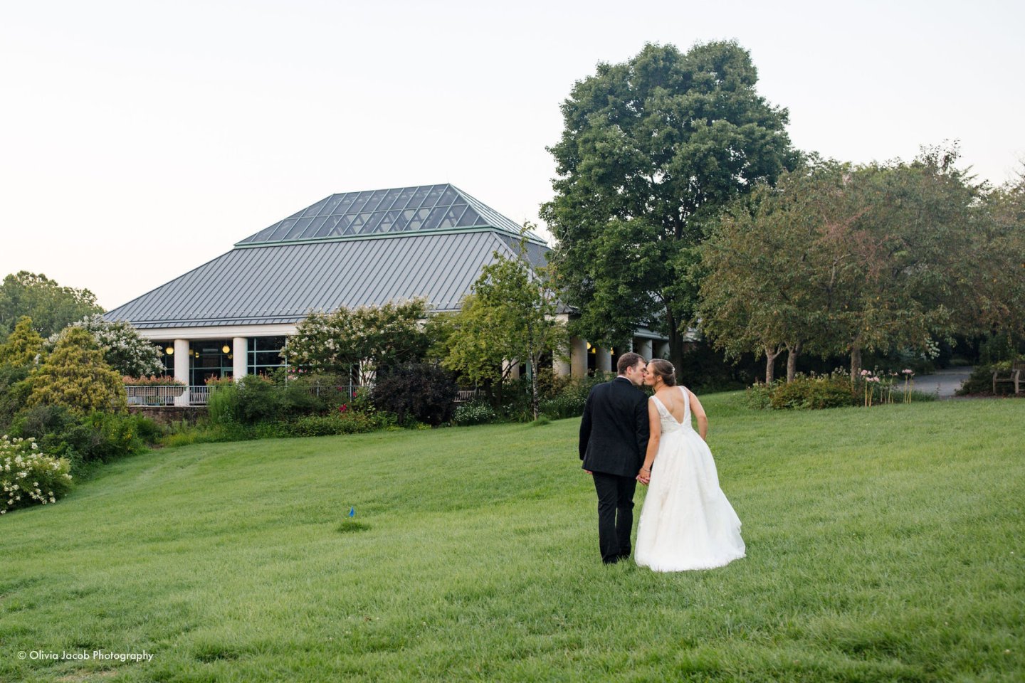 Couple in wedding attire walking on a grassy lawn towards a pavilion.